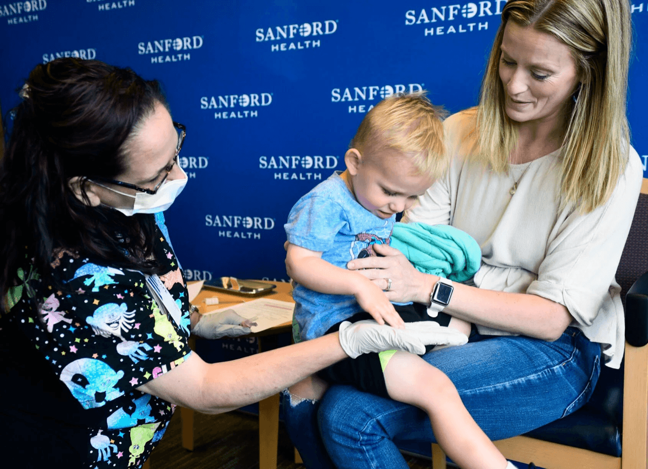 Memphis Lennick, one month shy of being 2 years old, gives a Sanford Health pediatric nurse a high-five after receiving a COVID-19 vaccination shot in his thigh while sitting on the lap of his mom, Nicole, at Sanford Health Bismarck north children's campus in June 2022.