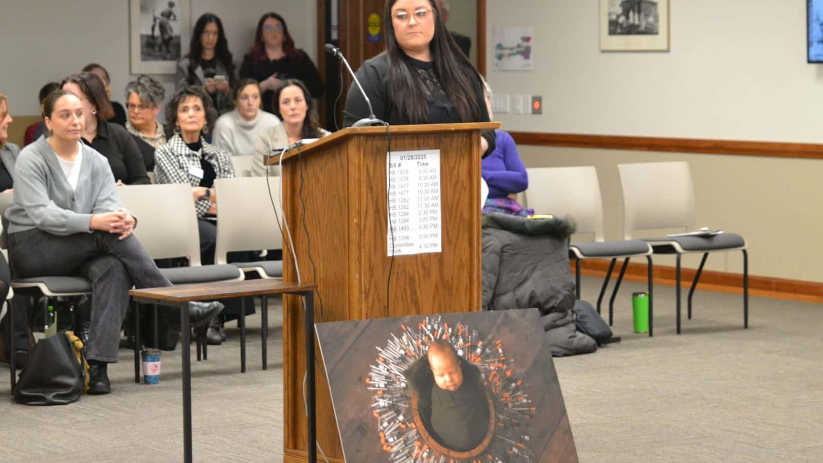 Abby Berger of Bismarck stands Jan. 29, 2025, before a legislative committee hearing with a photo illustrating the large number of syringes and vials needed to help her conceive her child. (Jeff Beach/North Dakota Monitor)