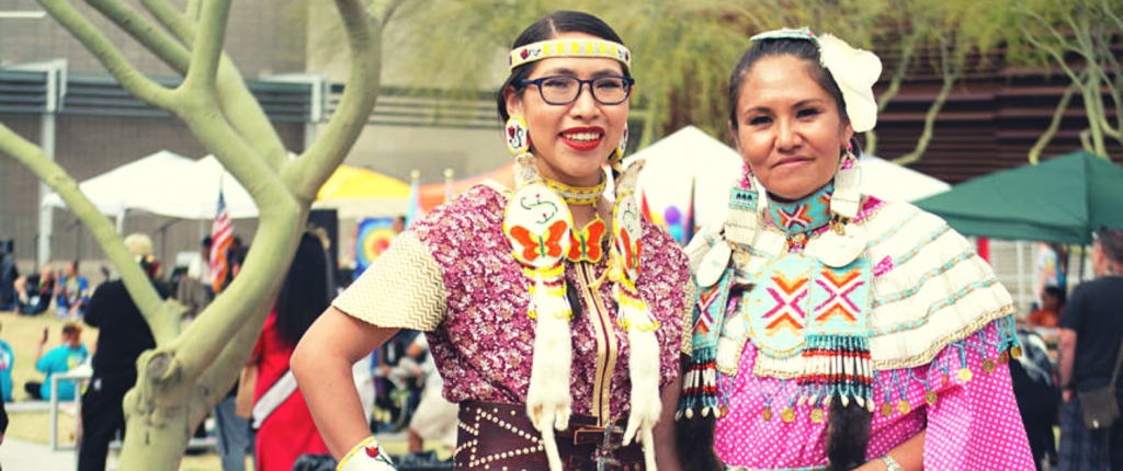 two native american women wearing traditional indigenous outfits at the two spirit powwow in Arizona with pink dress and wearing glasses