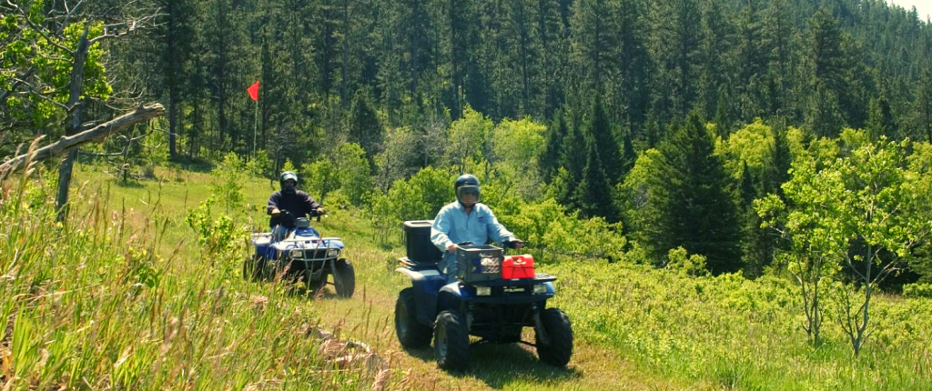 two men riding ATV vehicles in a forest wearing helmets and driving through grass