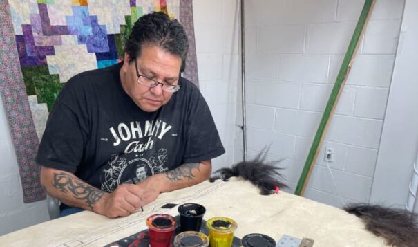 Native American artist Evans Flammond Sr. at work in the Whiteclay Makerspace in Whiteclay, Neb., applies paint to a buffalo robe that will eventually contain an elaborate encampment scene. Flammond is an established artist whose work appears in the South Dakota Capitol and who is now helping other artists explore their creativity and find a greater audience for their work. (Photo by Bart Pfankuch / South Dakota News Watch)